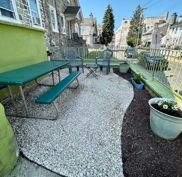 An outdoor patio with a table and chairs, surrounded by a gravel area and a metal fence.