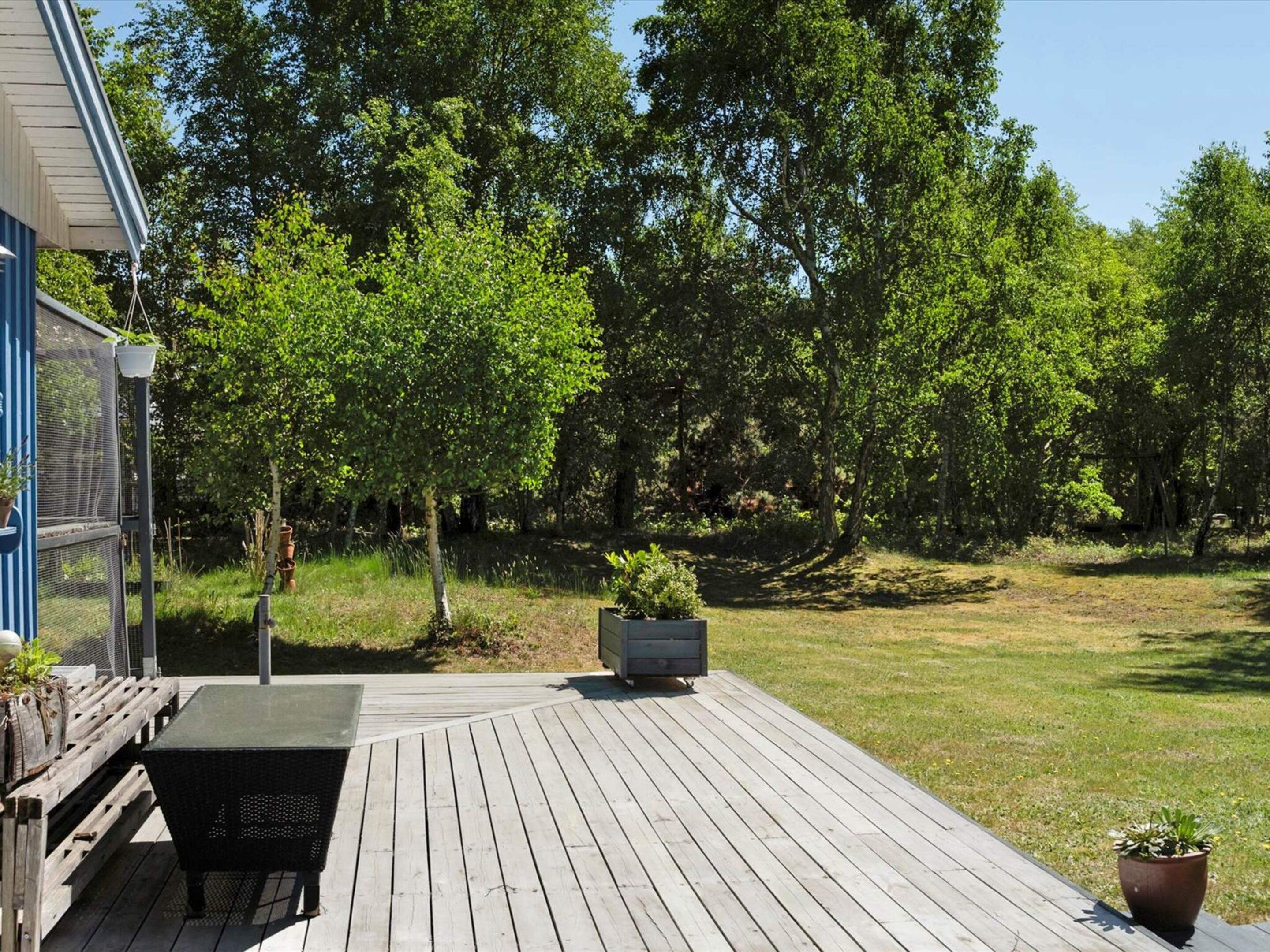 A wooden deck with a table and chairs, surrounded by greenery and trees.