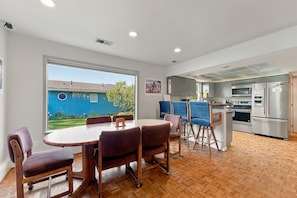 A modern kitchen with stainless steel appliances, a dining area with wooden furniture, and a view of a blue house outside. - The Red Lake House | Beach & Dock Access (Chelan)