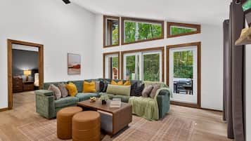 A spacious living room with a sectional sofa, ottomans, and a coffee table. There are large windows with multiple panes, a ceiling fan, and a view of greenery outside.