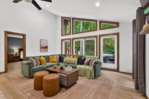 Ein geräumiges Wohnzimmer mit einem Ecksofa, Hocker und einem Couchtisch. Große Fenster mit vielen Sprossen, ein Deckenventilator und Blick ins Grüne sind ebenfalls vorhanden.
