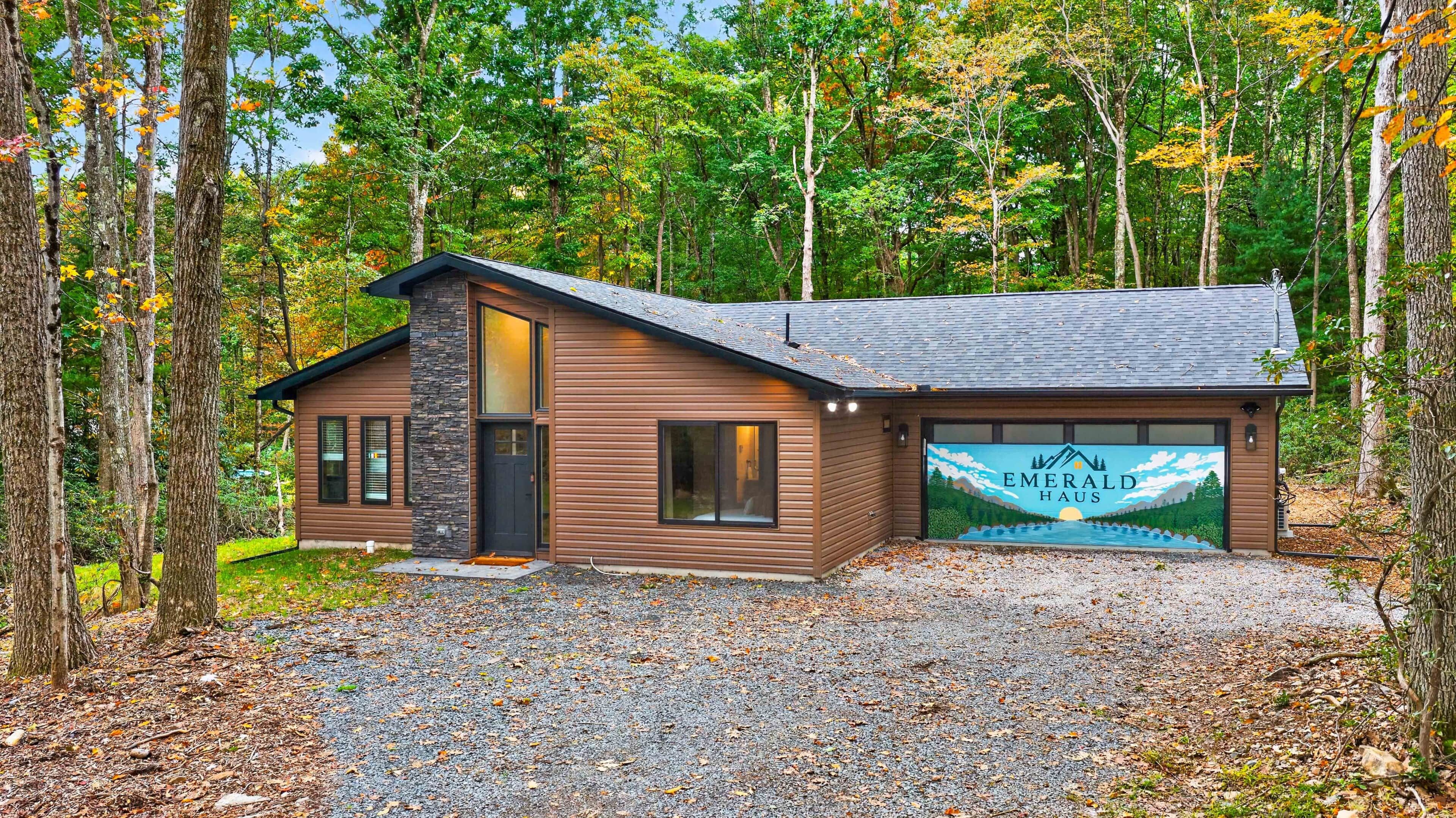 A cabin with a stone facade, a large garage door with a mural, and a gravel driveway surrounded by trees.