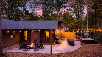 A wooden cabin with a fire pit and chairs, surrounded by trees and string lights.