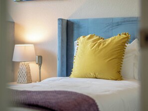 Una cama con edredón blanco y almohada amarilla, una lámpara de noche y un cabecero azul.