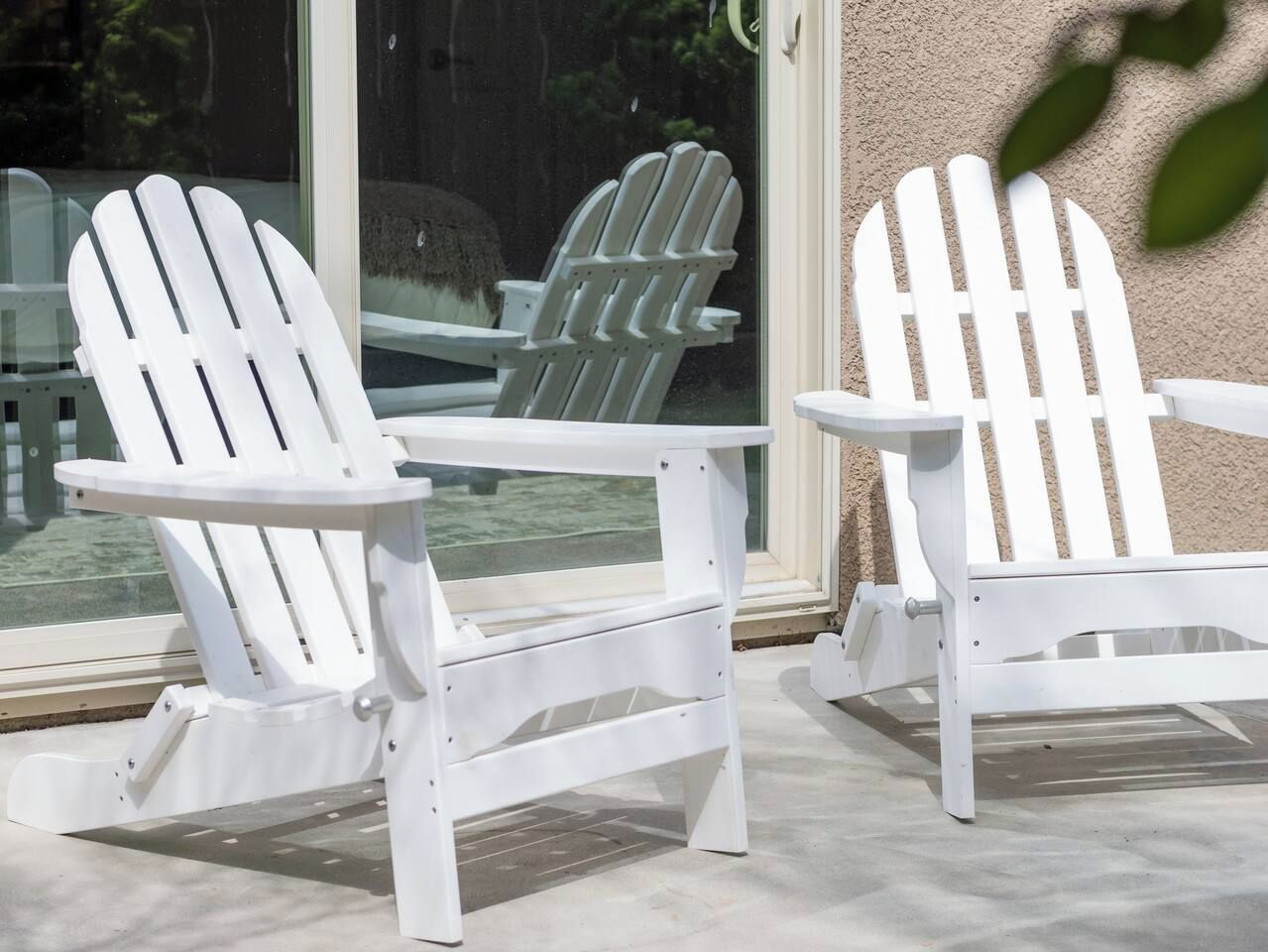 Two white Adirondack chairs on a patio with a glass door and a view of a garden.