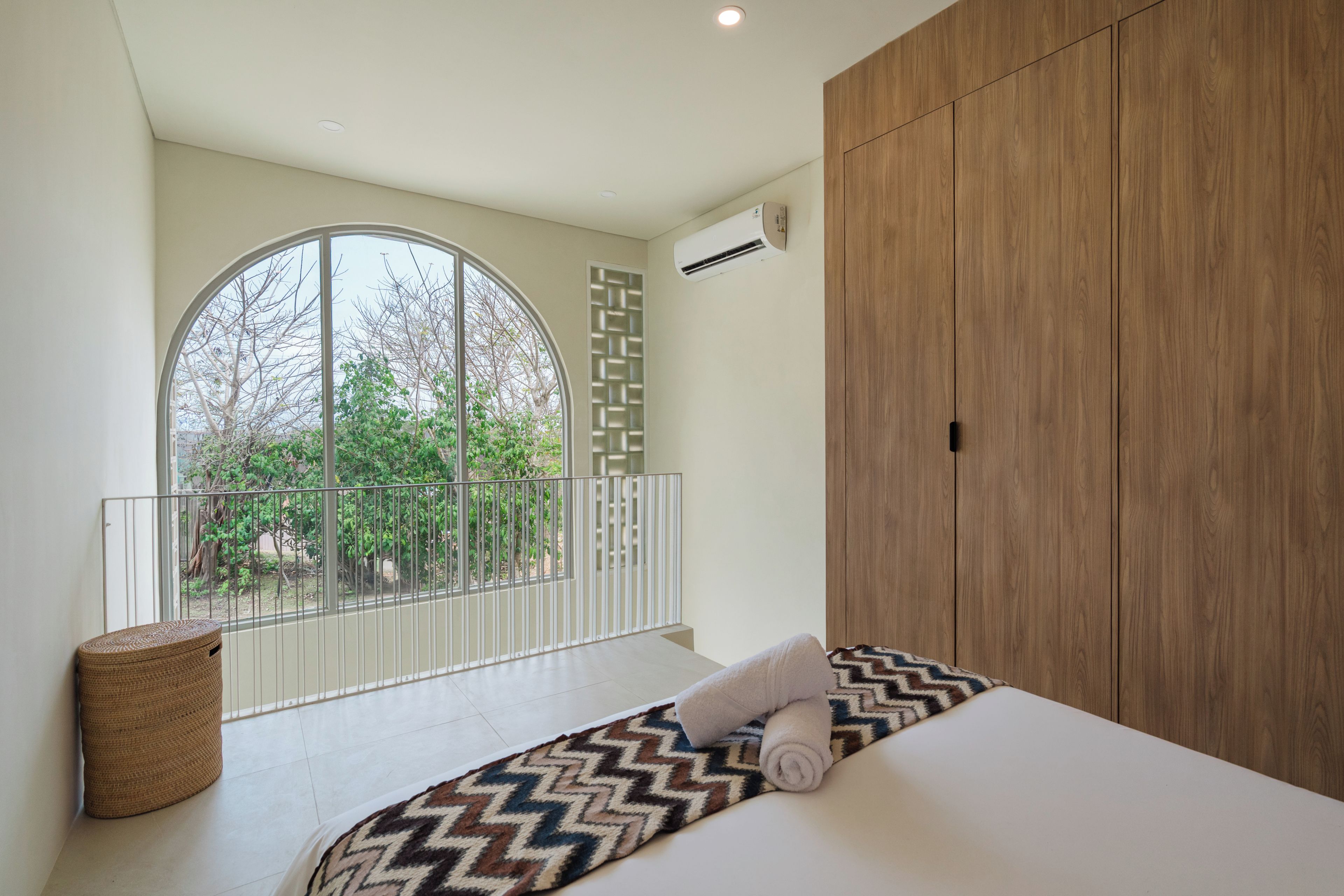 Un dormitorio con un ventanal grande, una cama con alfombra estampada, un armario de madera y una pared blanca.