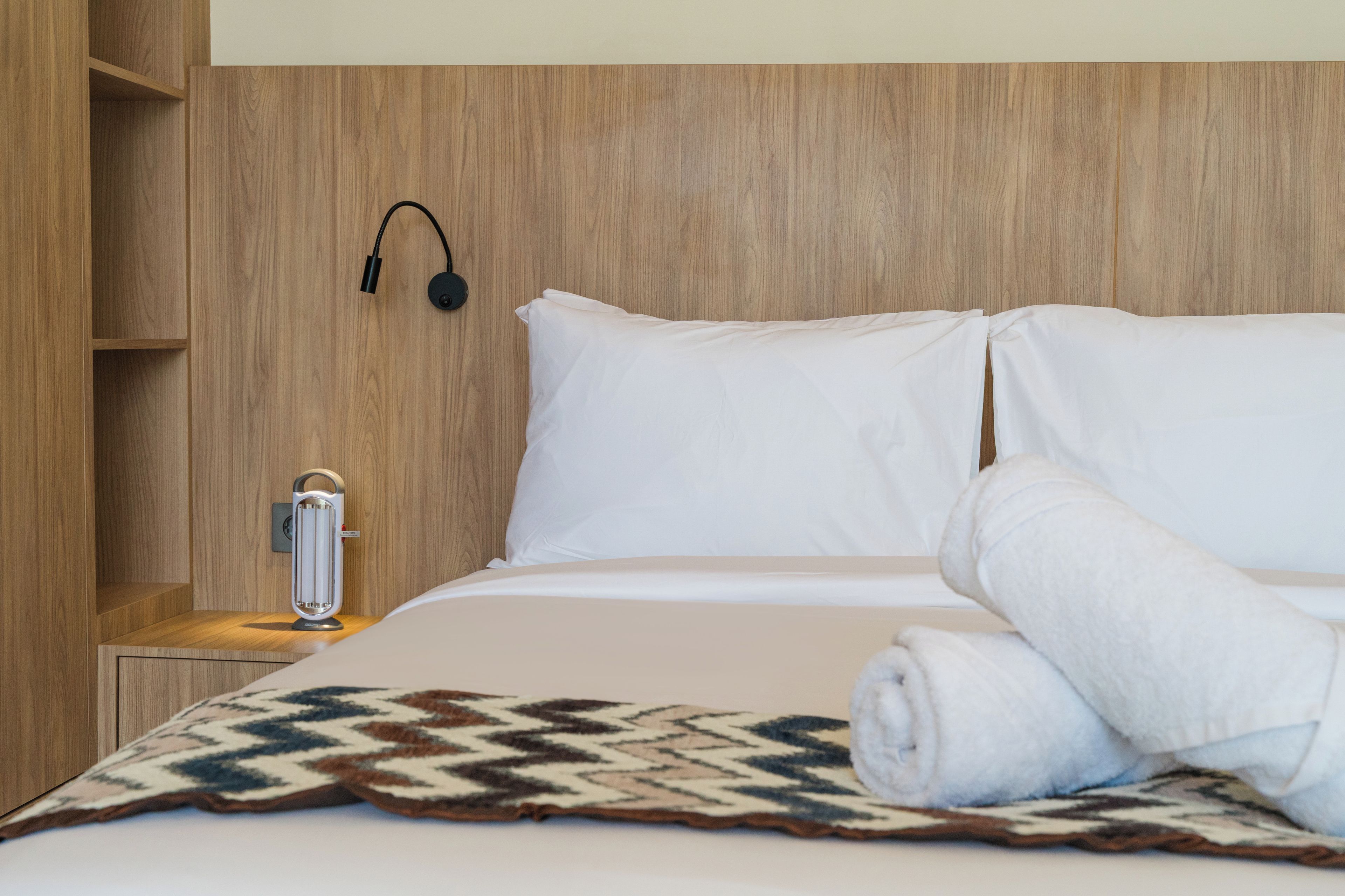Una habitación de hotel con cabecera de madera, cama con ropa de cama blanca, mesita de noche con lámpara y una manta.