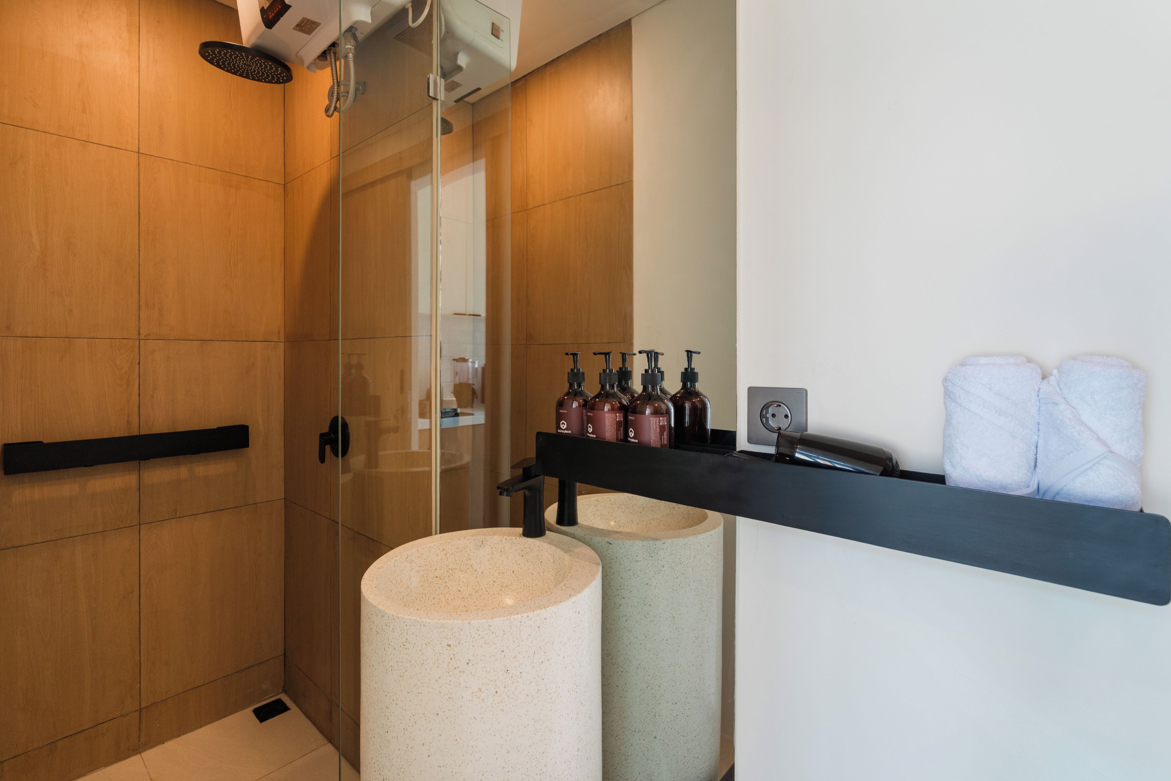 A modern bathroom with a glass shower enclosure, two round sinks, a black shelf with toiletries, and a towel rack.
