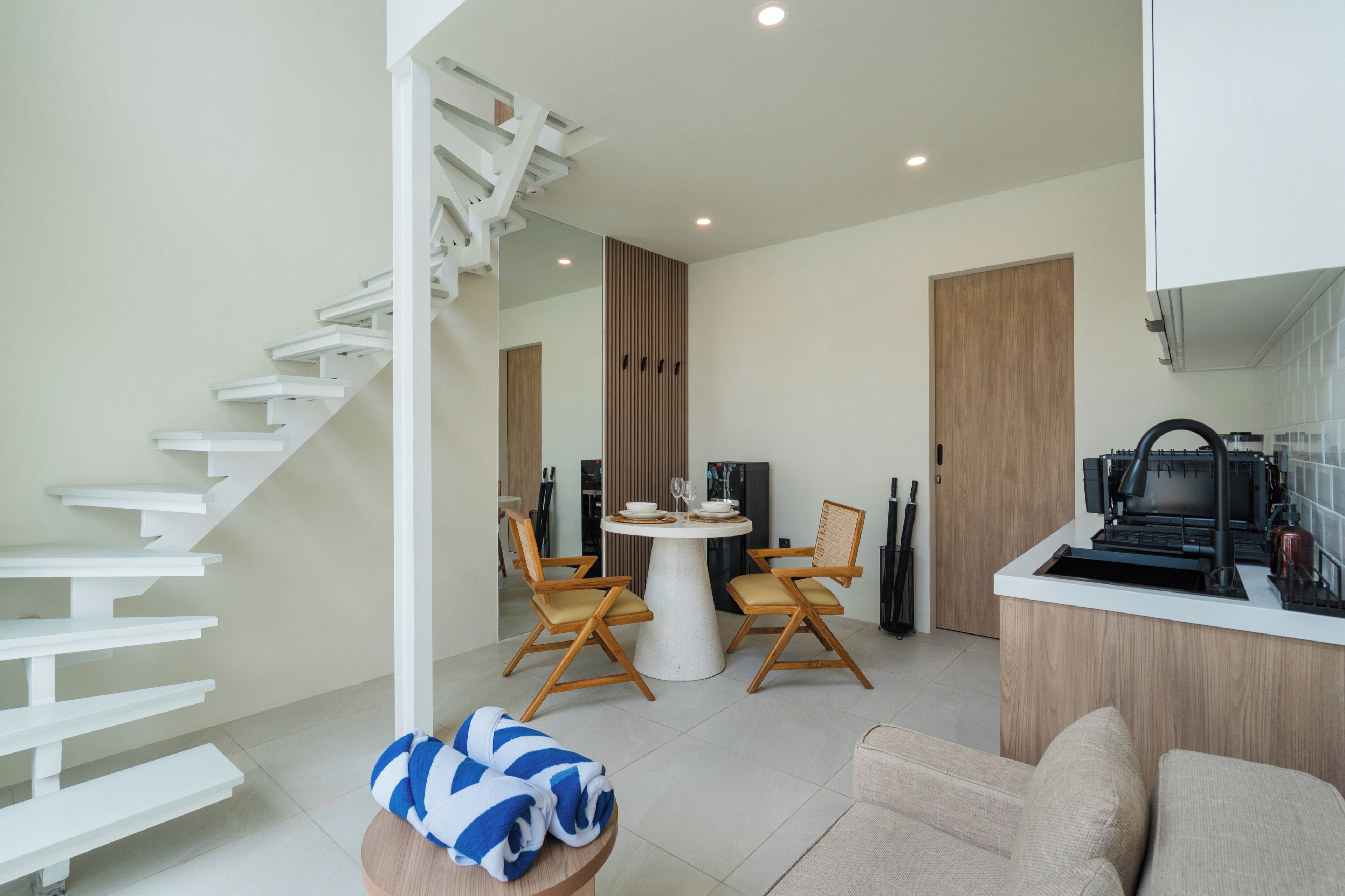 Een modern interieur met een witte trap, een eethoek met twee houten stoelen, een ronde tafel en een kitchenette met een koffieapparaat.
