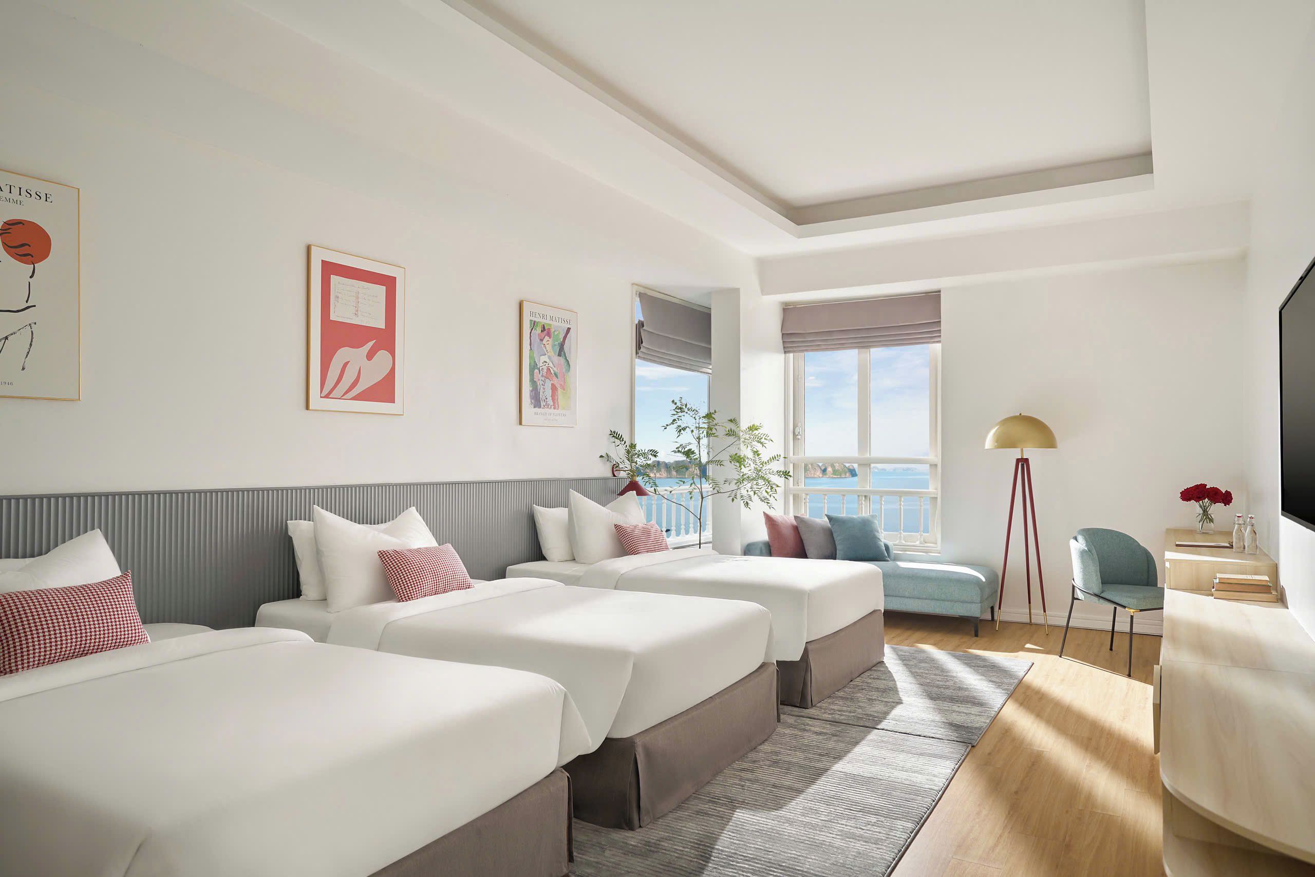 Une chambre d’hôtel avec deux lits, un bureau, une chaise, une fenêtre donnant sur un beau paysage et des œuvres d’art accrochées aux murs.