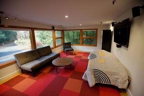 Ein Zimmer mit einer Couch, einem Sessel, einem kleinen Tisch und einem Bett mit gemustertem Überwurf. Große Fenster bieten Blick auf Grünflächen außerhalb.