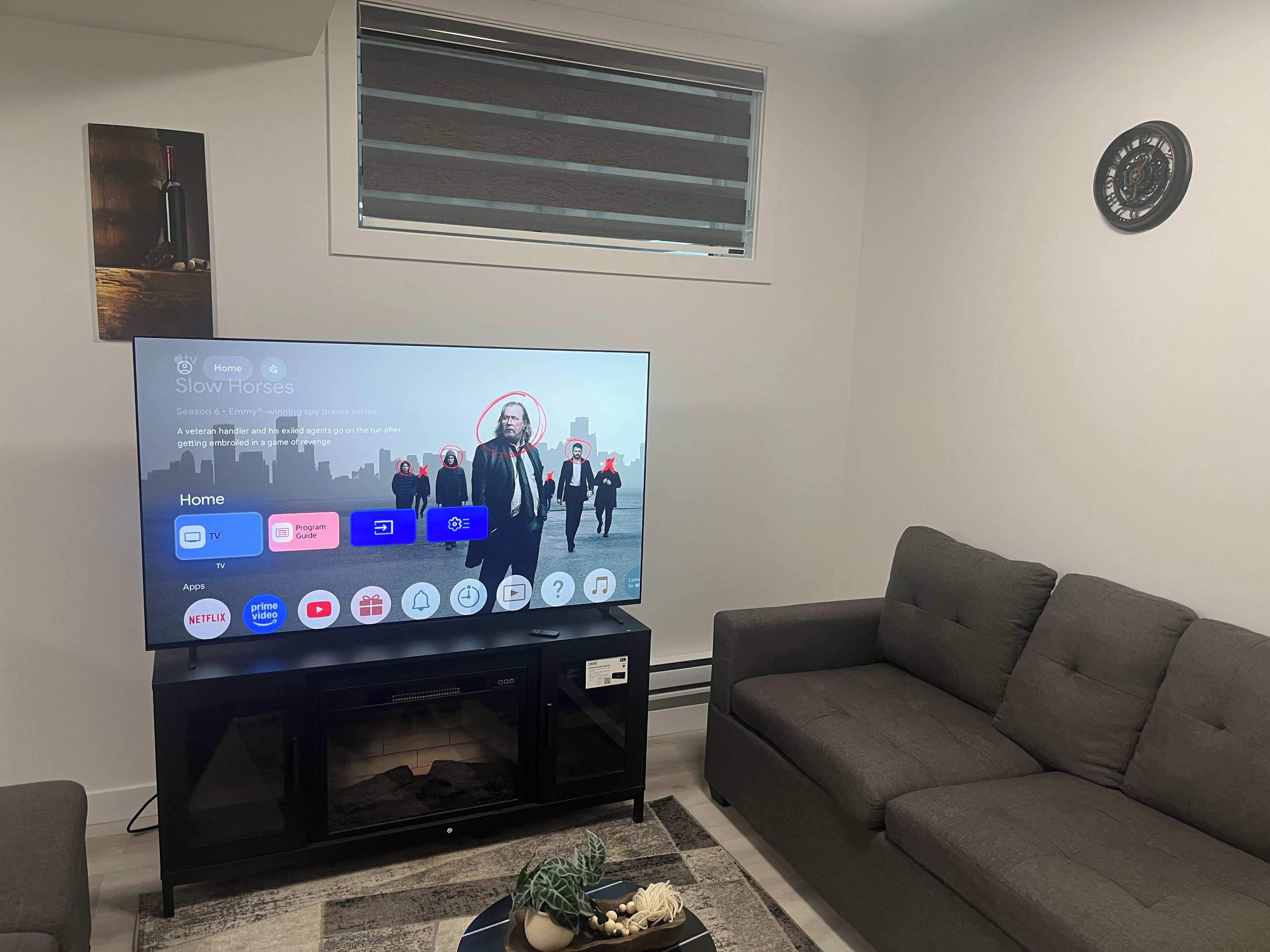 Ein Wohnzimmer mit einem Flachbildfernseher auf einem Schrank, einem grauen Sofa, einem kleinen Tisch mit einer Pflanze und einem Wandbild.