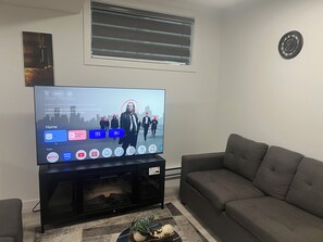 Ein Wohnzimmer mit einem Flachbildfernseher auf einem Schrank, einem grauen Sofa, einem kleinen Tisch mit einer Pflanze und einem Wandbild.