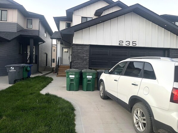 A residential driveway with a white SUV parked, a garage with the number 235, and two green waste bins.