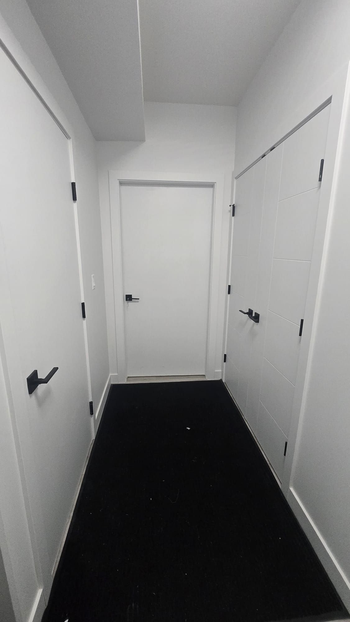 Un couloir étroit aux murs blancs et au sol noir, qui mène à une porte fermée au bout.