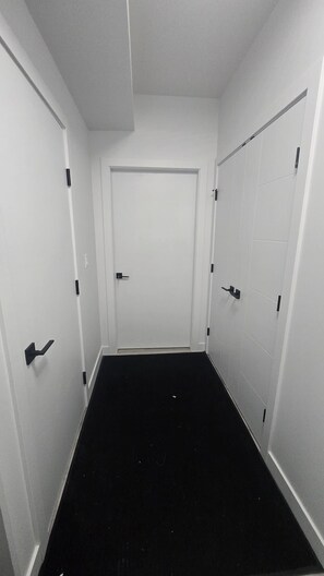 A narrow hallway with white walls and black flooring, leading to a closed door at the end.