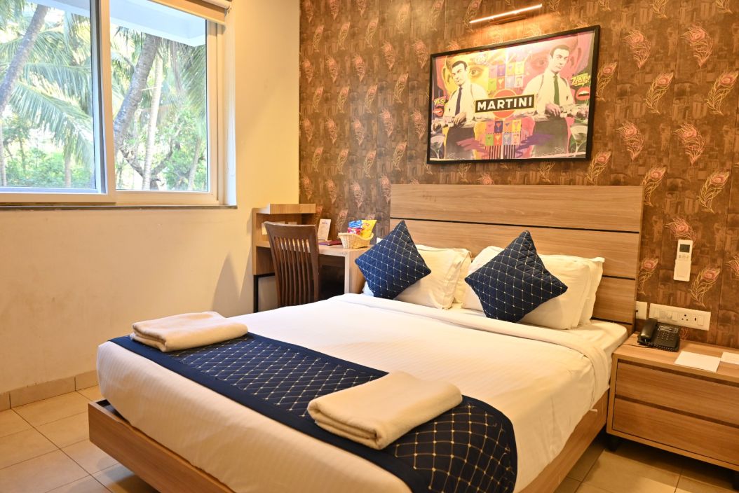 Habitación de hotel con cama, escritorio, silla, televisor y ventana con vista a palmeras.
