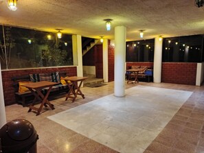 Amplo espaço interno com parede de tijolos, um banco de madeira e uma mesa. Há colunas e uma escadaria ao fundo.