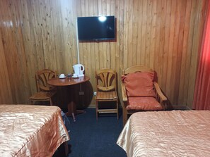 Quarto com duas camas, televisão na parede, mesa com chaleira e xícaras, e duas cadeiras.