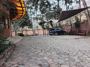 A paved courtyard with a car parked under a canopy, surrounded by trees and a brick wall.