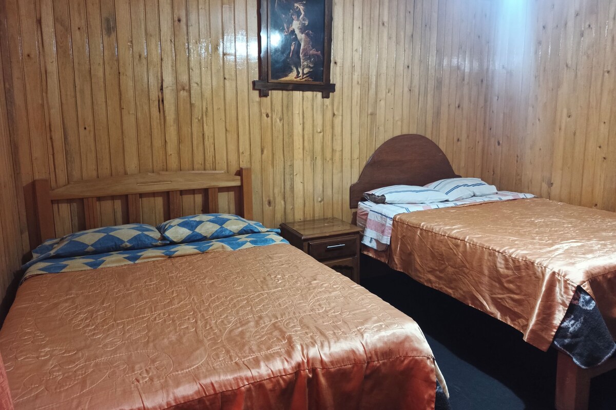 Quarto com duas camas de madeira, um banco de madeira, uma mesa de cabeceira e um quadro emoldurado na parede.