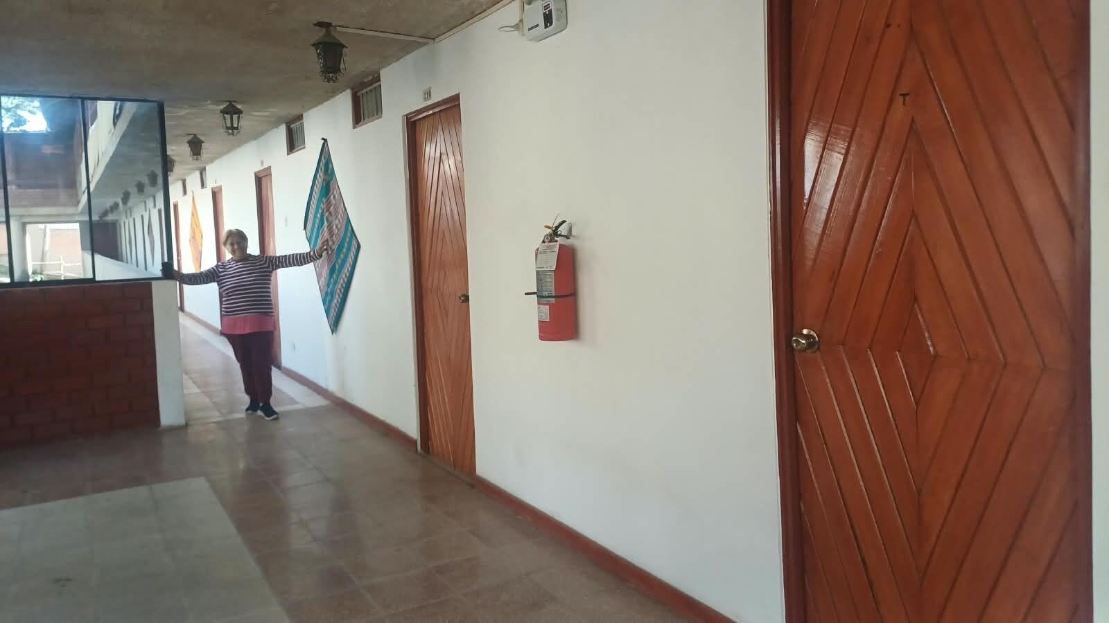 A hallway with a fire extinguisher mounted on the wall, a wooden door on the right, and a person standing near the end.
