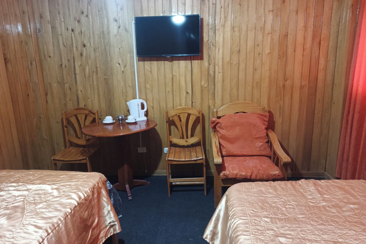 A room with two beds, a television mounted on the wall, a table with a kettle and cups, and two chairs.