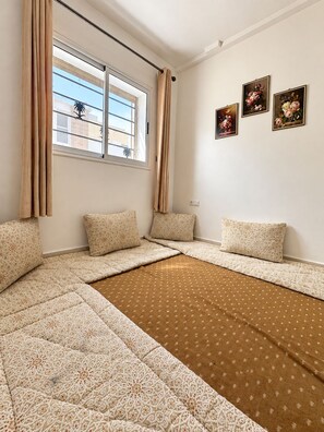 A room with a large bed, beige patterned bedding, a window with curtains, and three framed floral paintings on the wall. - Between Tradition and Modernity — Premium Concierge Service (Dakhla)