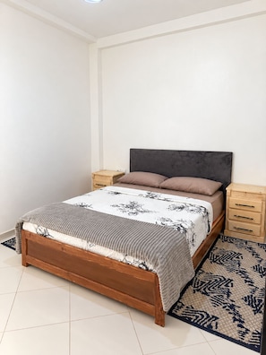 A bedroom with a wooden bed, a dark headboard, a patterned bedspread, two bedside tables, and a patterned rug.