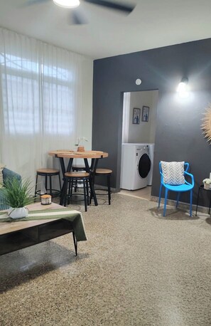 Ein Wohnzimmer mit einem runden Tisch, Stühlen, einem blauen Sessel, einer Waschmaschine und einem Deckenventilator.