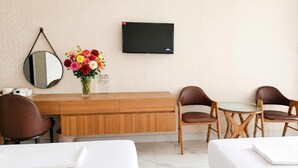 Ein Hotelzimmer mit einem Holztisch, zwei braunen Ledersesseln, einem kleinen Tisch, einem an der Wand montierten Fernseher, einem runden Spiegel und einer Vase mit bunten Blumen.