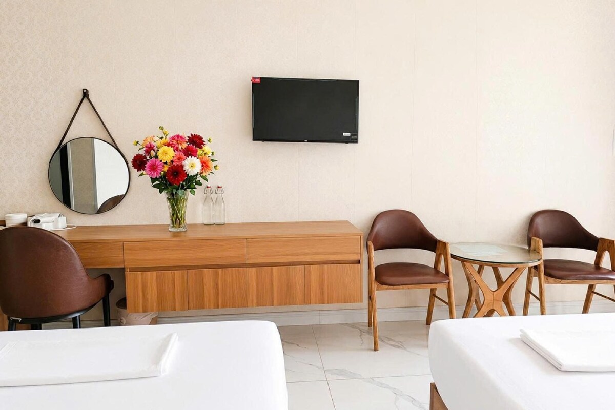 Ein Hotelzimmer mit einem Holztisch, zwei braunen Ledersesseln, einem kleinen Tisch, einem an der Wand montierten Fernseher, einem runden Spiegel und einer Vase mit bunten Blumen.