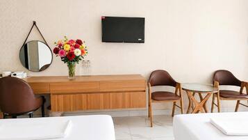Habitación de hotel con un escritorio de madera, dos sillones de cuero marrón, una mesita, un televisor montado en la pared, un espejo redondo y un florero con flores coloridas.