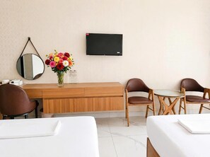 Ein Hotelzimmer mit einem Holztisch, zwei braunen Ledersesseln, einem kleinen Tisch, einem an der Wand montierten Fernseher, einem runden Spiegel und einer Vase mit bunten Blumen.