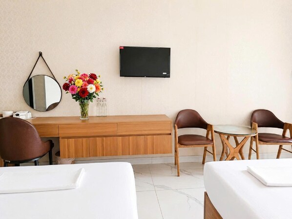 A hotel room with a wooden desk, two brown leather chairs, a small table, a TV mounted on the wall, a round mirror, and a vase with colorful flowers.