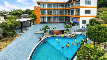 A swimming pool with people around it, a building with orange and white exterior, and a clear sky.