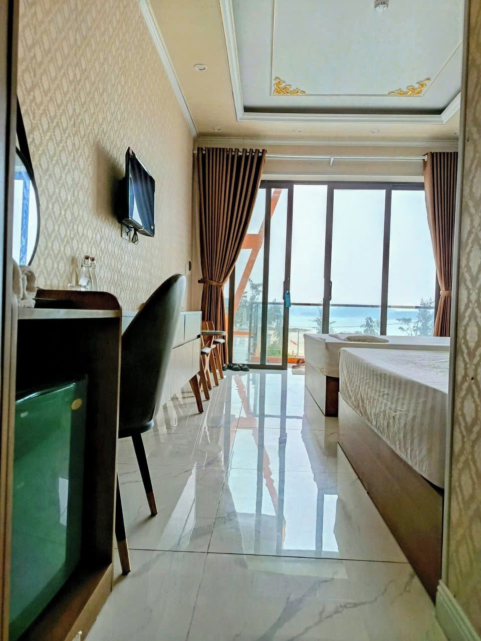 Quarto de hotel com cama, escrivaninha, cadeira, televisão e uma grande janela com vista para o mar.