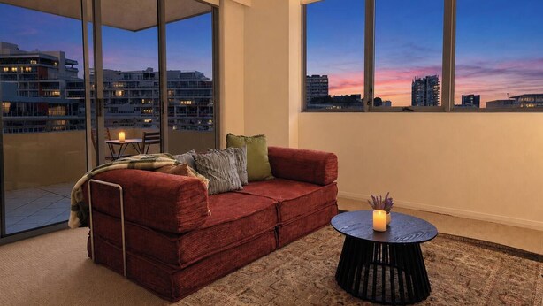 A modern living room with a red sofa, a round black coffee table, and a view of the city skyline at dusk. - The Ember | 2BR|2BA|Pool, Gym, Sauna + Parking | Boutique Stay near Sydney CBD (Waterloo)