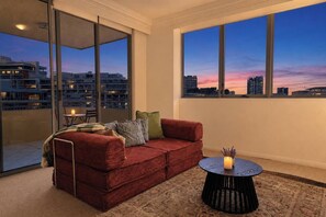 A modern living room with a red sofa, a round black coffee table, and a view of the city skyline at dusk. - The Ember | 2BR|2BA|Pool, Gym, Sauna + Parking | Boutique Stay near Sydney CBD (Waterloo)