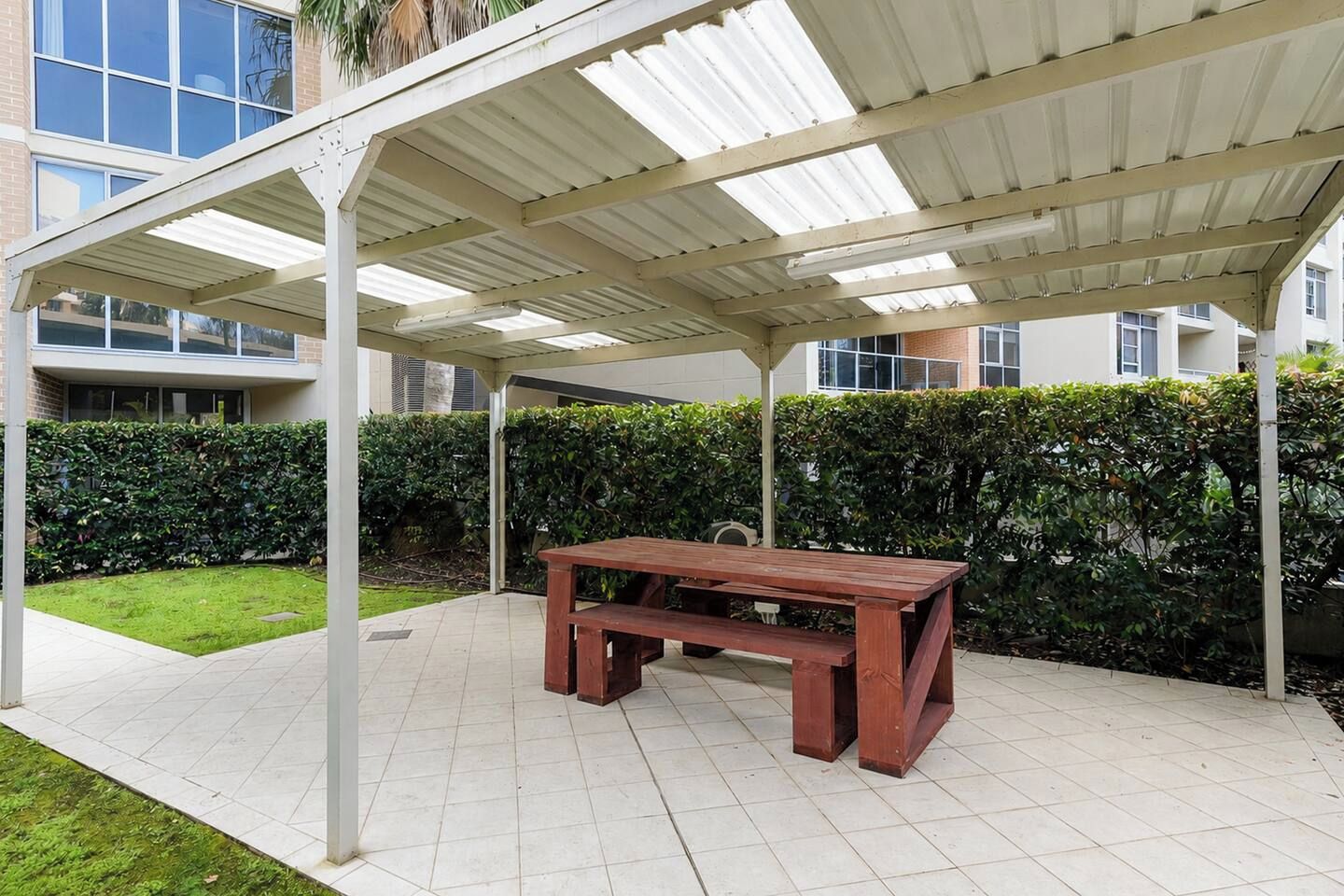 A covered outdoor area with a wooden bench, a tiled floor, and a hedge in the background.
