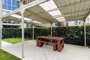 A covered outdoor area with a wooden bench, a tiled floor, and a hedge in the background. - The Ember | 2BR|2BA|Pool, Gym, Sauna + Parking | Boutique Stay near Sydney CBD (Waterloo)