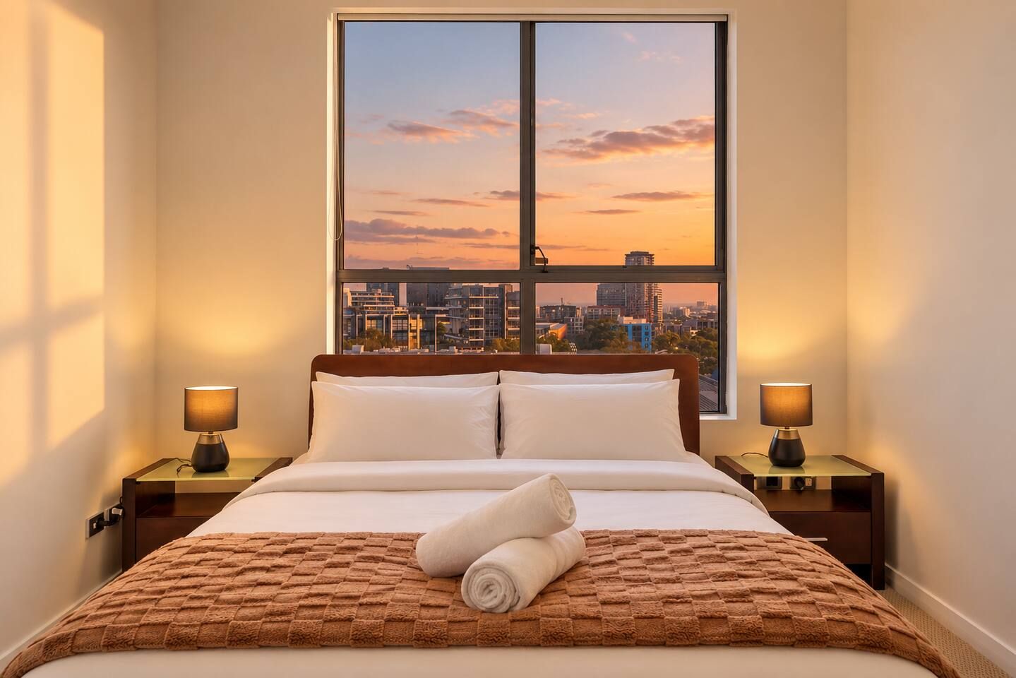 A hotel room with a bed, two bedside lamps, a window with a city view, and a rolled towel on the bed.