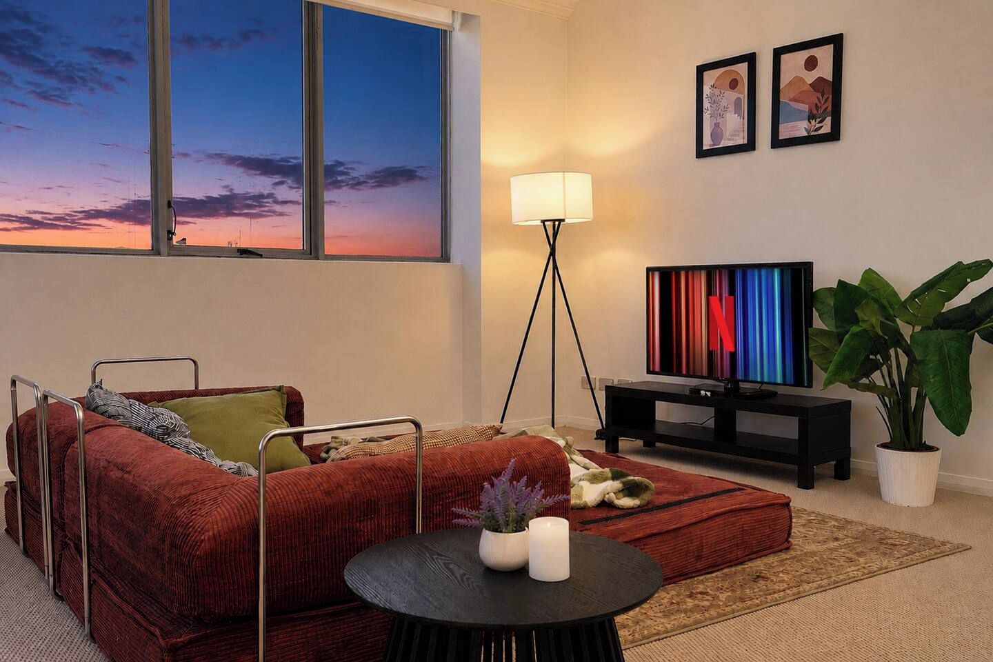 A modern living room with a red sofa, a small round table with candles, a TV on a stand, and a large window with a sunset view.