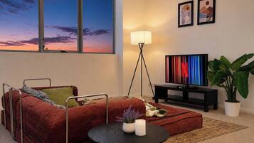 A modern living room with a red sofa, a small round table with candles, a TV on a stand, and a large window with a sunset view.