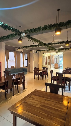 A dining area with wooden tables and chairs, hanging pendant lights, and a view into the kitchen area.