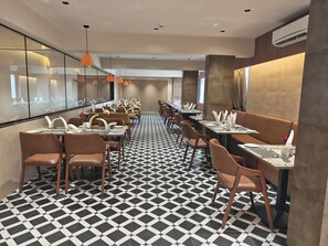 Ein Restaurant mit einem schwarz-weißen Schachbrettmuster auf dem Boden, mehreren Tischen mit weißen Servietten und orangefarbenen Pendelleuchten.