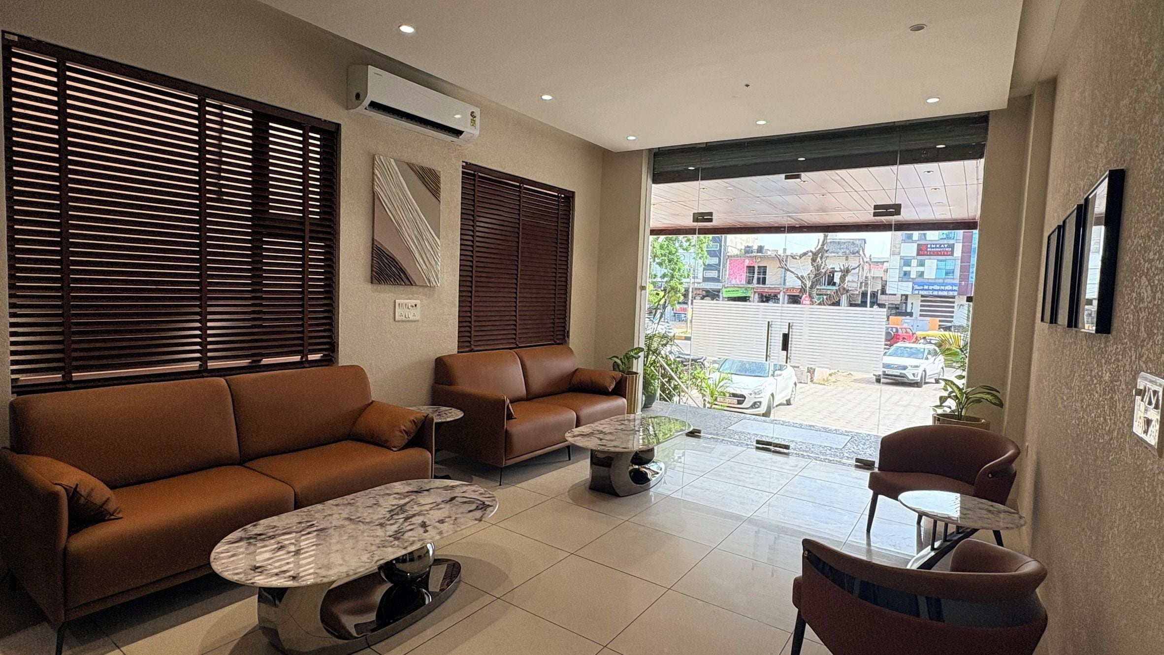 A modern living room with a brown sofa, a coffee table, and a chair. There are two windows with blinds, an air conditioning unit, and a view of the street outside.