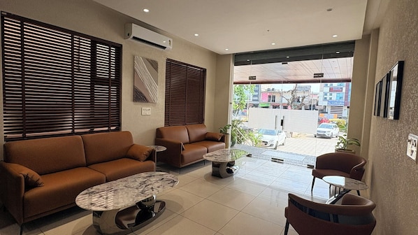 A modern living room with a brown sofa, a coffee table, and a chair. There are two windows with blinds, an air conditioning unit, and a view of the street outside.