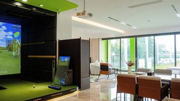 A modern hotel room with a large screen displaying a golf game, a seating area with a table and chairs, and a view of the outdoors through large windows.