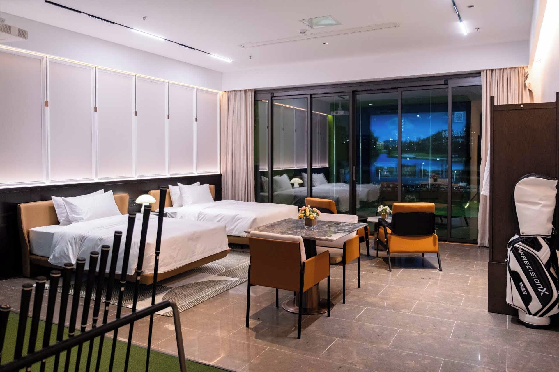 Habitación de hotel con dos camas, una mesa de comedor, sillas, un bolso de golf y un amplio ventanal con vistas.